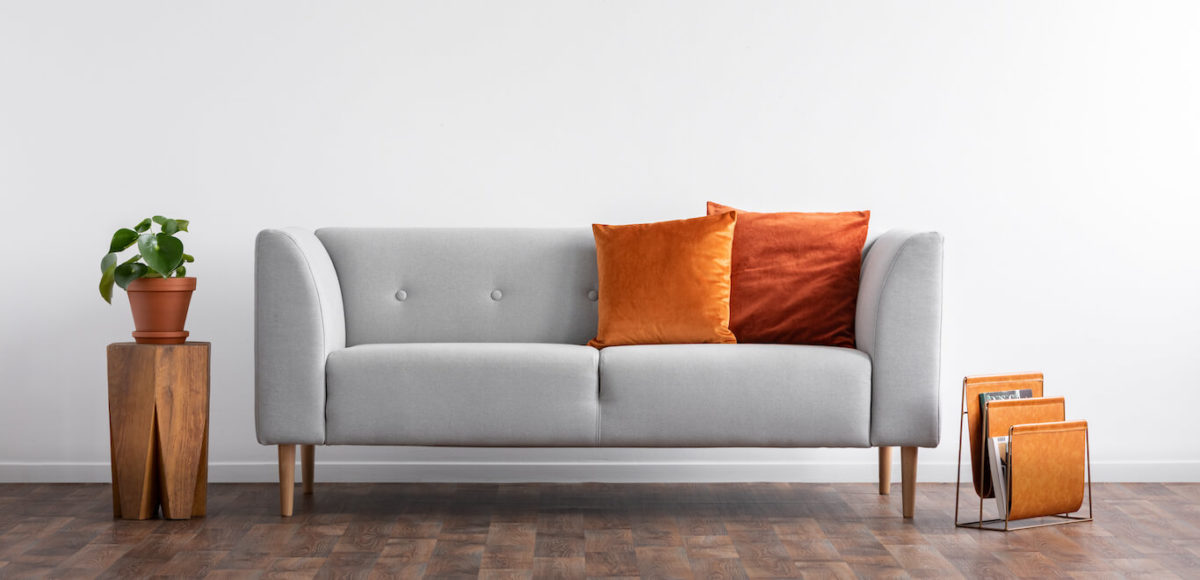 A modern couch in light gray with burnt orange pillows sits in front of blank white wall.