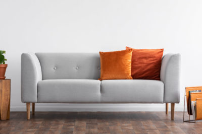 A modern couch in light gray with burnt orange pillows sits in front of blank white wall.
