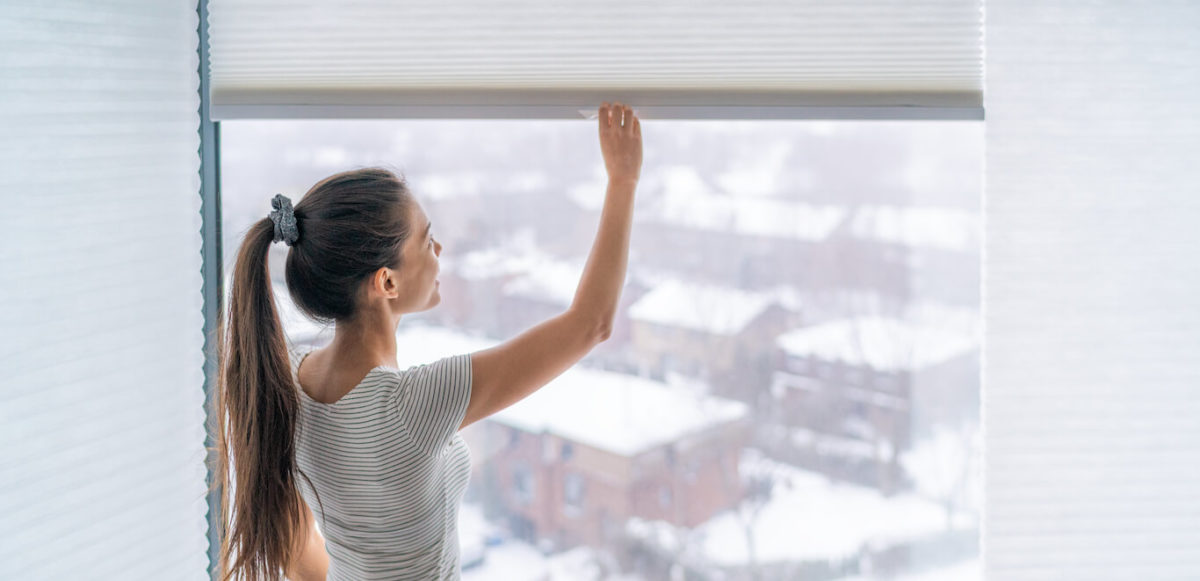 Young woman with ponytail pulls down the window shades with snow visible outdoors.