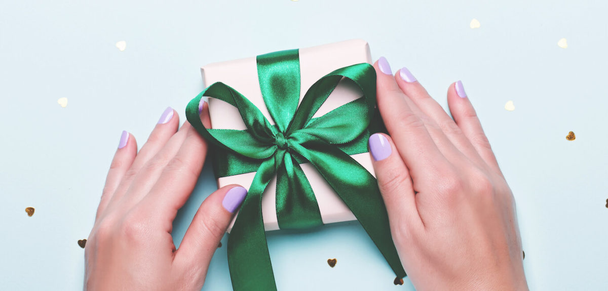 A woman's hands arrange a small gift wrapped in a satin green bow on a light blue background.