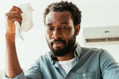 Sweating man holding napkin with focus on broken air conditioner behind him.