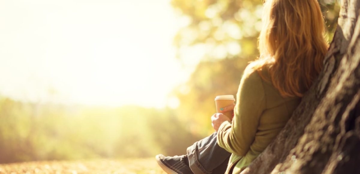 Blonde woman sitting alone in front of tree trunk in autumn with a paper coffee cup in hand.