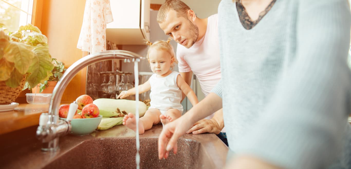 Parents and infant child in kitchen observing running faucet water.