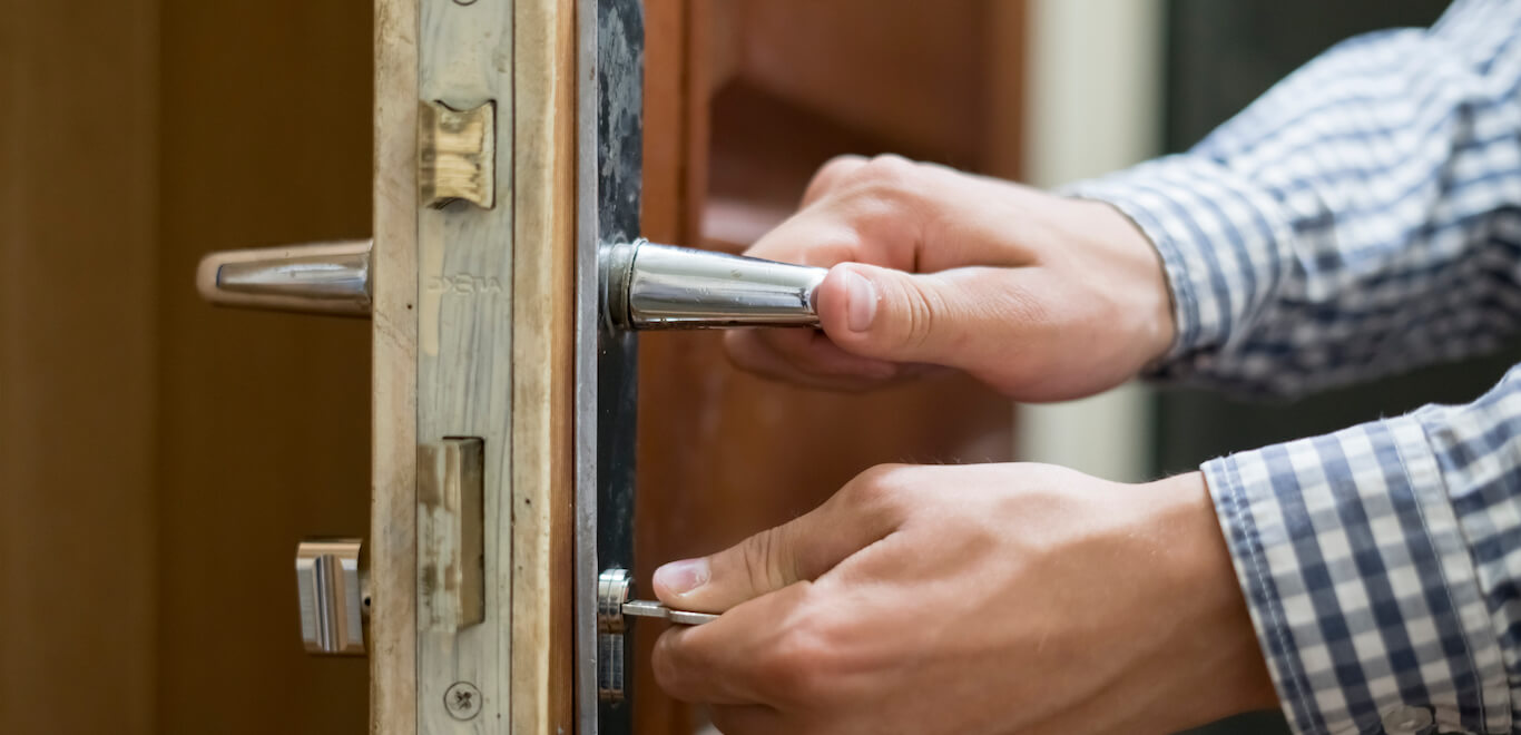 A man in a checkered shirt unlocks a door while twisting the handle to enter.