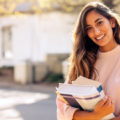 Young woman in sweatshirt is smiling and holding books on college campus.