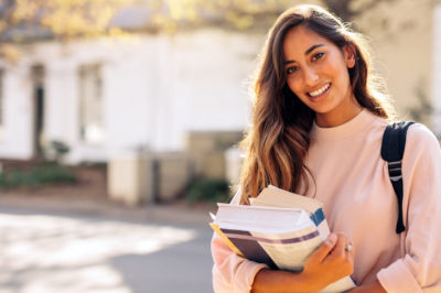 Young woman in sweatshirt is smiling and holding books on college campus.