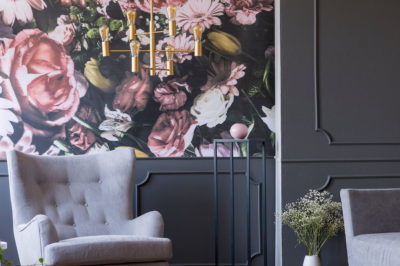 Grey armchairs and a funky gold chandelier against a bold floral wallpaper in a maximalist apartment.