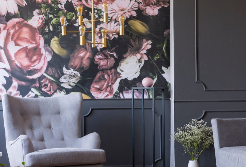 Grey armchairs and a funky gold chandelier against a bold floral wallpaper in a maximalist apartment.