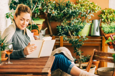 A woman enjoying a glass of wine and reading a book on a patio she has made more private using plants and greenery.