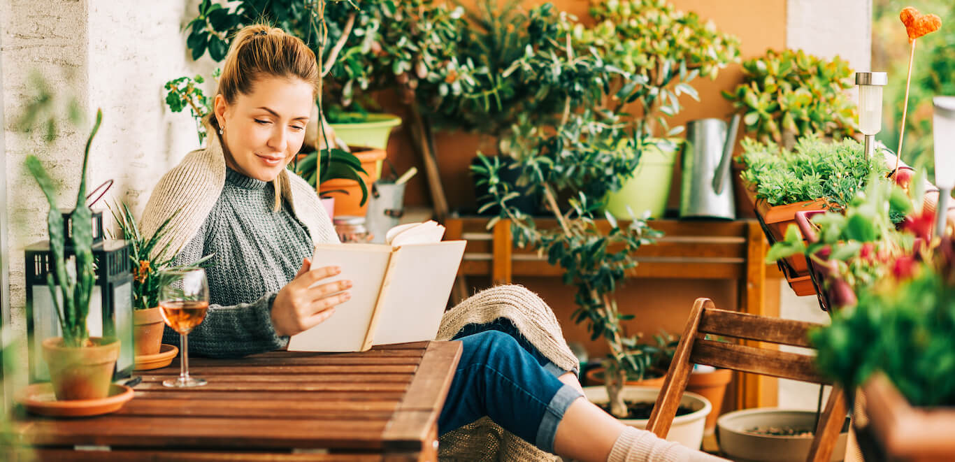 A woman enjoying a glass of wine and reading a book on a patio she has made more private using plants and greenery.