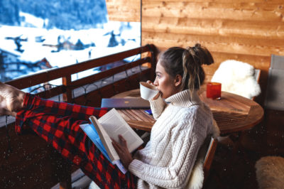 Woman on balcony in winter enjoying a book and cup of coffee.