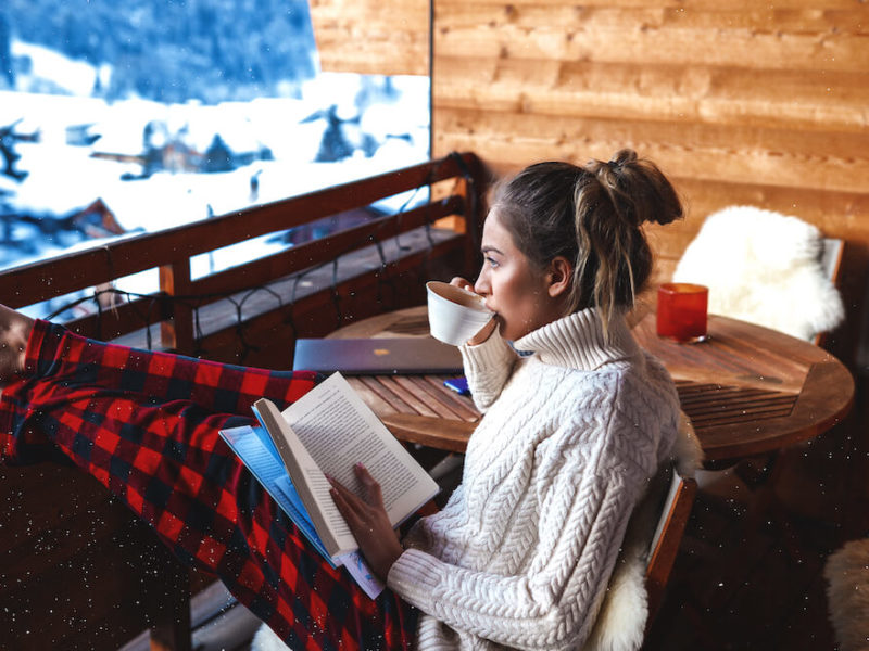 Woman on balcony in winter enjoying a book and cup of coffee.
