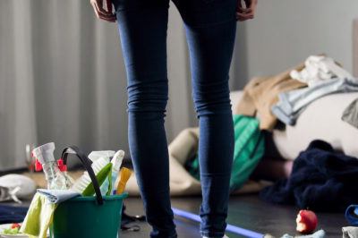Woman stands in apartment looking at mess of clothes and old food surrounded by cleaning supplies ready to use apartment cleaning ideas.