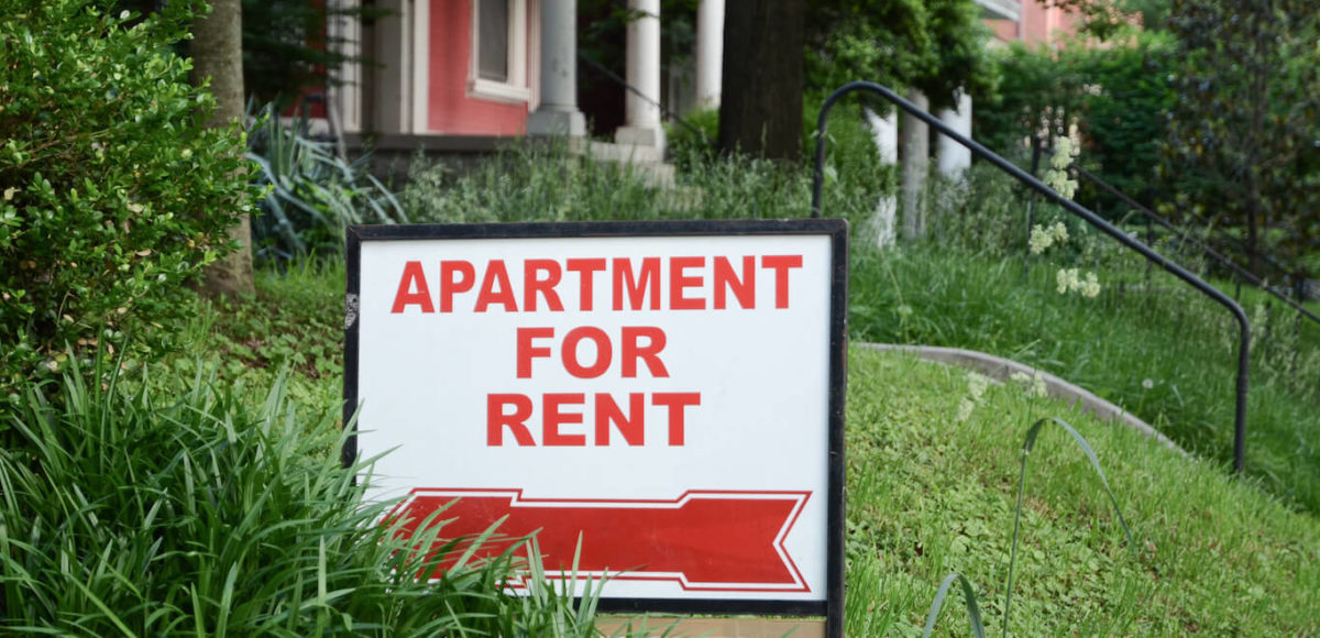 Sign outside of walk up brick apartment that says Apartment For Rent in a lush green lawn.