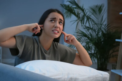 A young woman with her fingers in her ears, looking up towards loud noise outside her apartment.