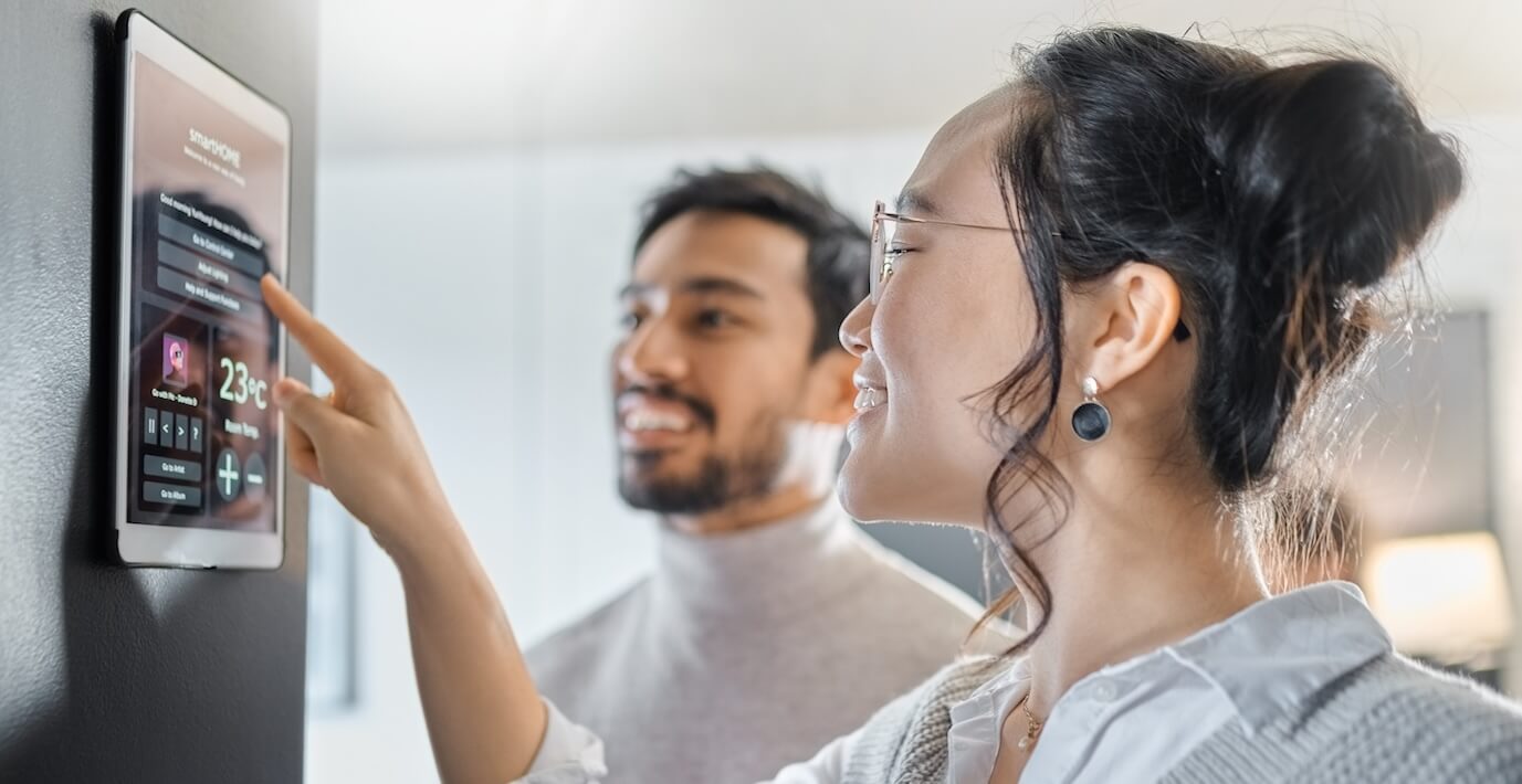 A young couple uses a tablet on the wall to adjust the temperature of their apartment.